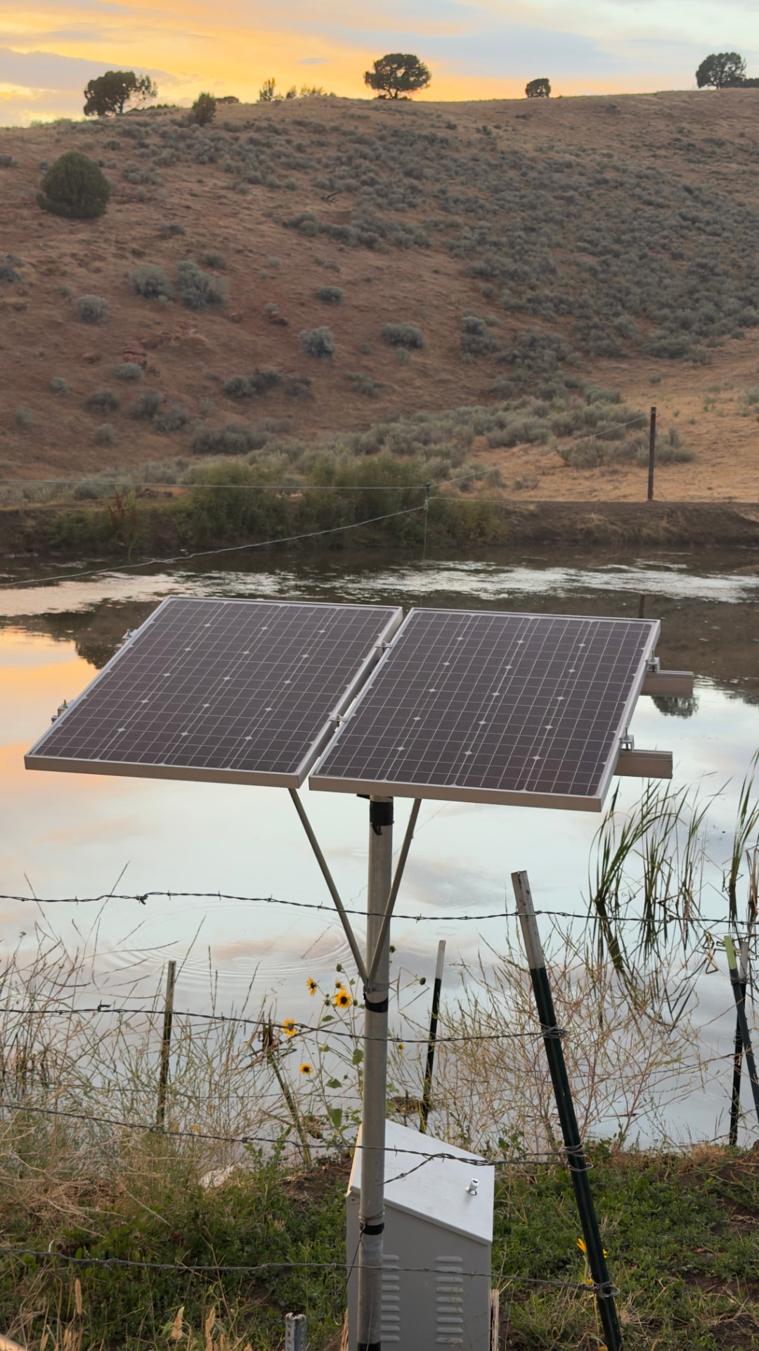 Solar panel array on a pole at the edge of a ranch pond at sunset